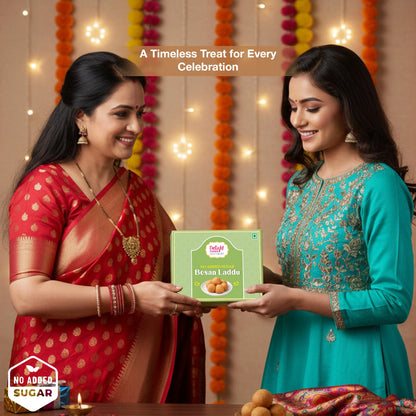 Two women exchanging a box of Besan Laddu with decorative lights and flowers in the background.